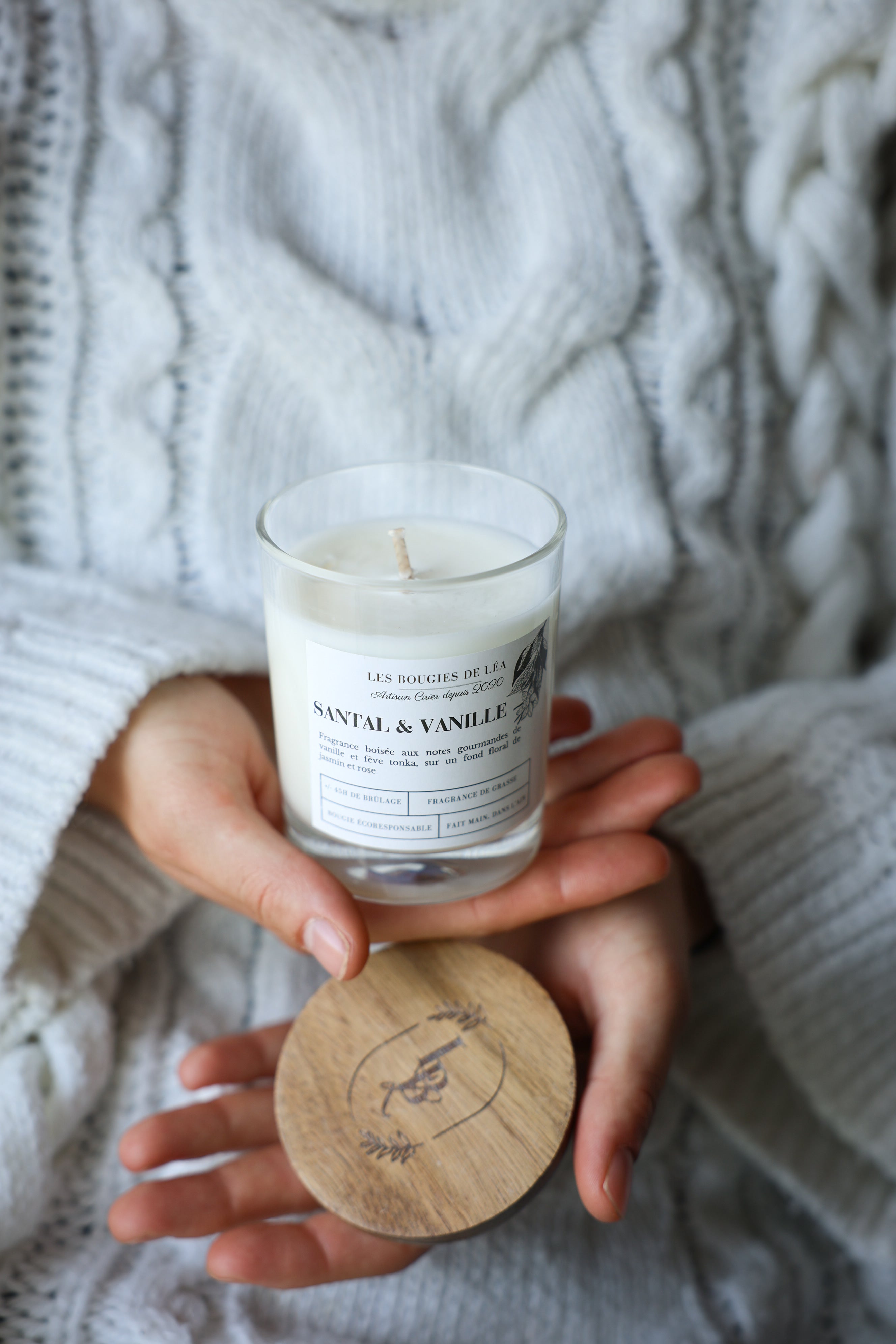 Une femme portant un pull en laine ou coton blanc, tenant dans ses mains une Bougie artisanale parfumée Les Bougies de Léa Santal & Vanille avec un couvercle en bois d'Acacia.