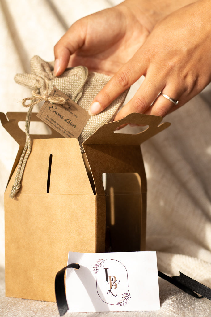 Coffret Cadeaux créations artisanales parfumées Les Bougies de Léa LBL. Une main sortant un pochon en toile de jute d'un coffret en carton et carte de visite LBL.