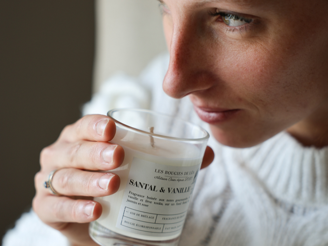 Léa la créatrice Les Bougies de Léa - LBL tenant dans sa main une bougie parfumée Santal & Vanille pour la sentir, habillée d'un pull en laine blanc.