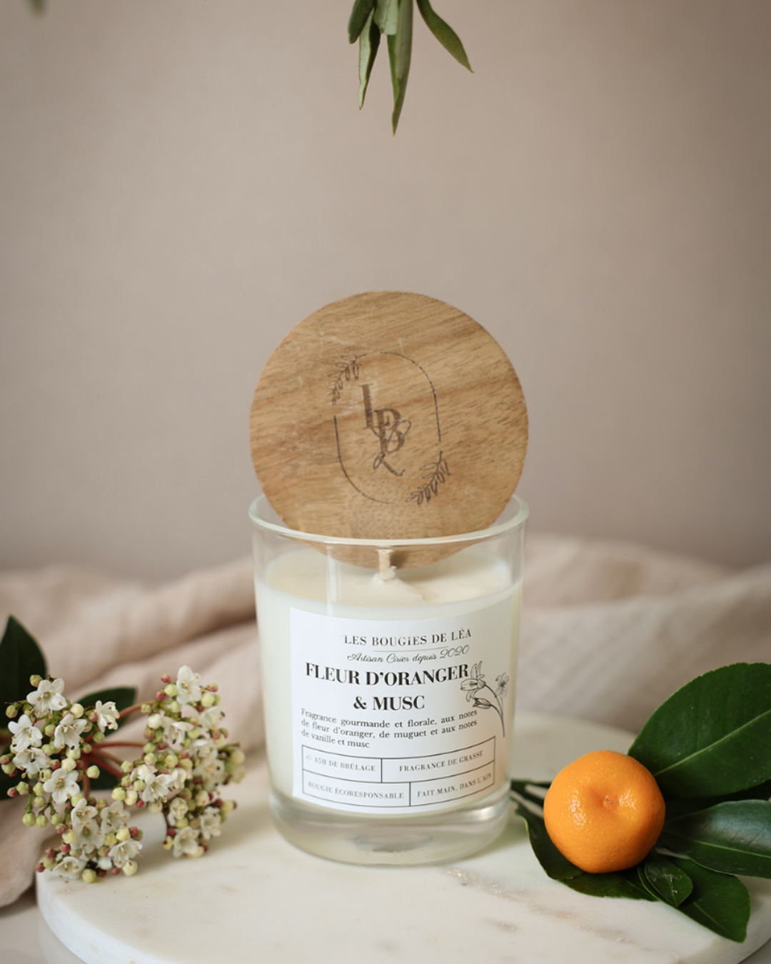 Bougie artisanale parfumée Fleur d'oranger & Musc Les Bougies de Léa avec couvercle en bois d'acacia, sur un support en marbre blanc, avec un drap en lin en arrière plan, accompagnée d'une orange et des fleurs blanches.