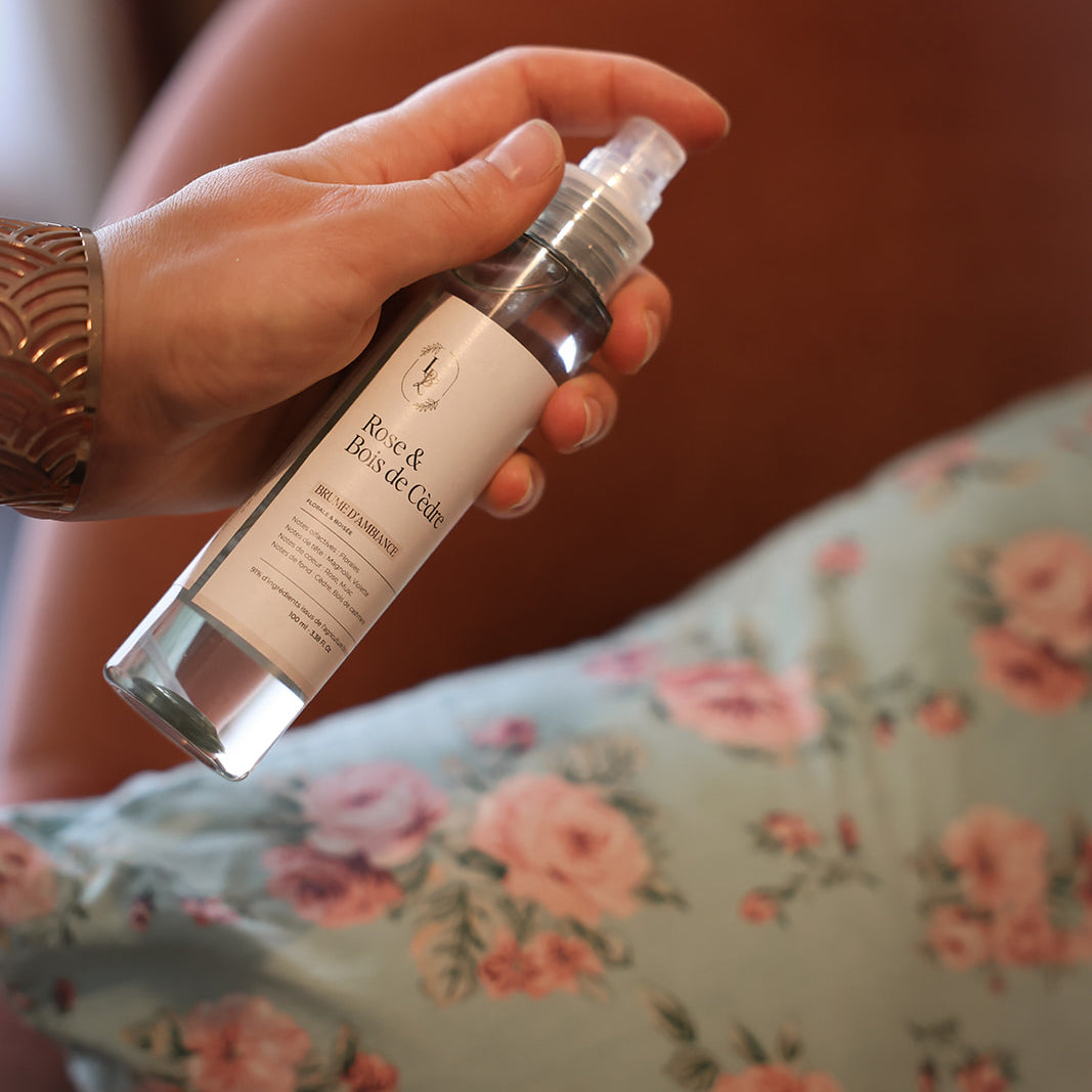 Main pulvérisant de la brume d'ambiance parfumée LBL Rose & Bois de Cèdre sur une coussin aux motifs fleuris.