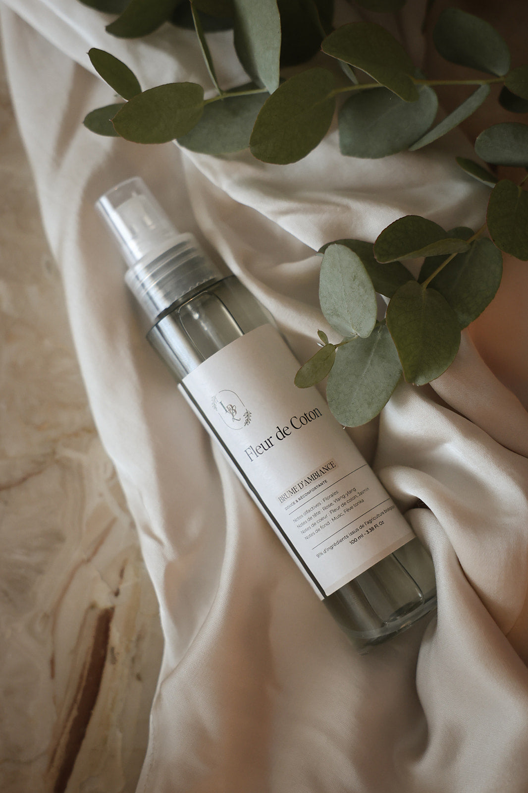 Testeur de brume d'ambiance LBL 30ml parfum Fleur de Coton posé sur un drapé en tissu beige avec des feuilles d'eucalyptus.