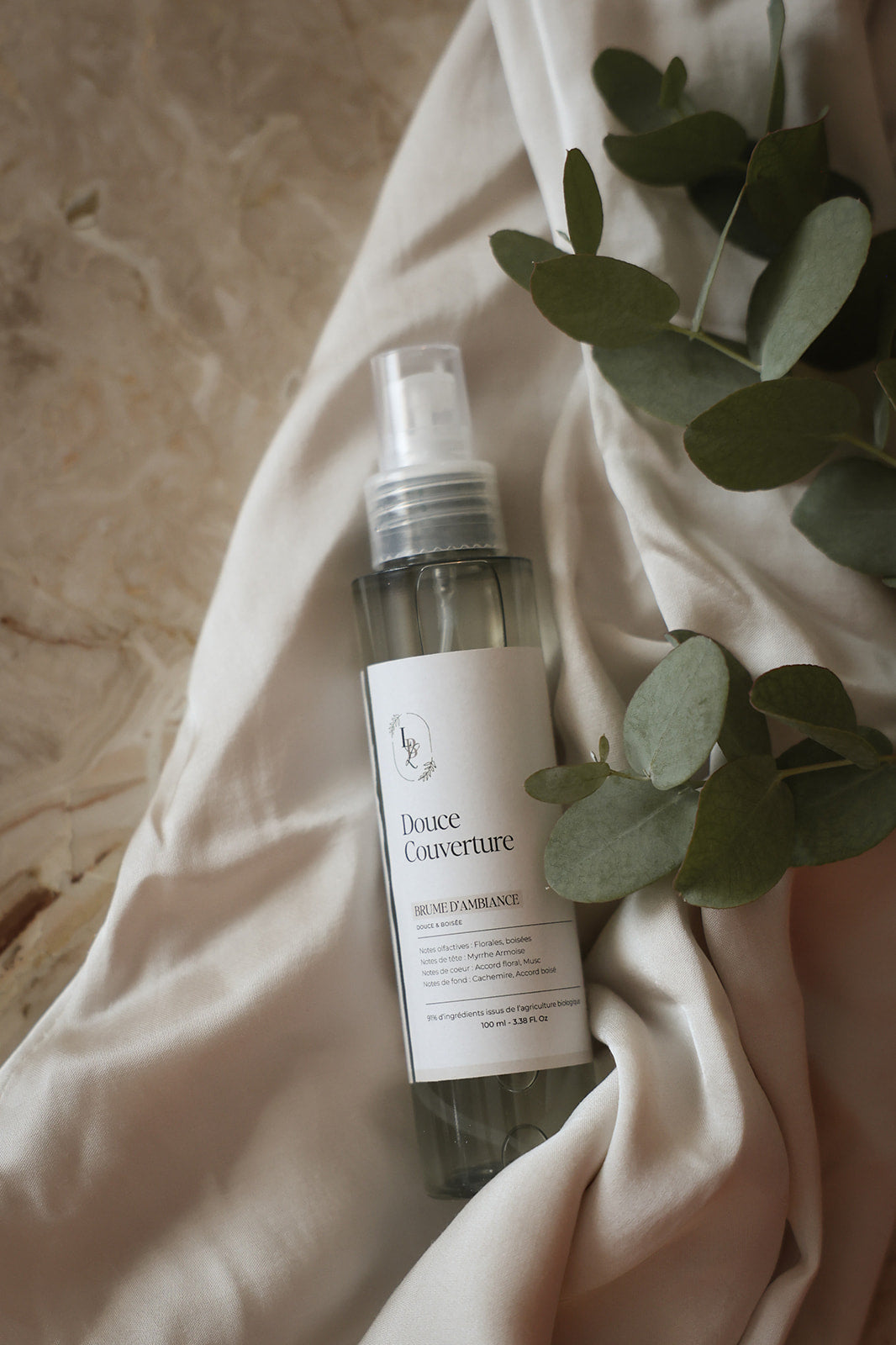 Brume d'ambiance parfumée Douce couverture LBL sur un drapé blanc satinéet feuille d'eucalyptus.