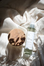 Brume d'ambiance Douce couverture LBL sur un drapé blanc satiné avec rayon du soleil et ombres végétales et florales, à côté d'une bougie parfumée Les Bougies de Léa avec le couvercle en bois d'acacia.