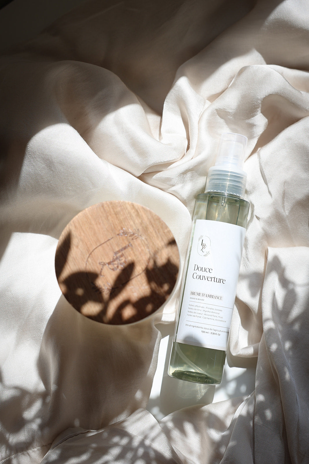 Brume d'ambiance Douce couverture LBL sur un drapé blanc satiné avec rayon du soleil et ombres végétales et florales, à côté d'une bougie parfumée Les Bougies de Léa avec le couvercle en bois d'acacia.