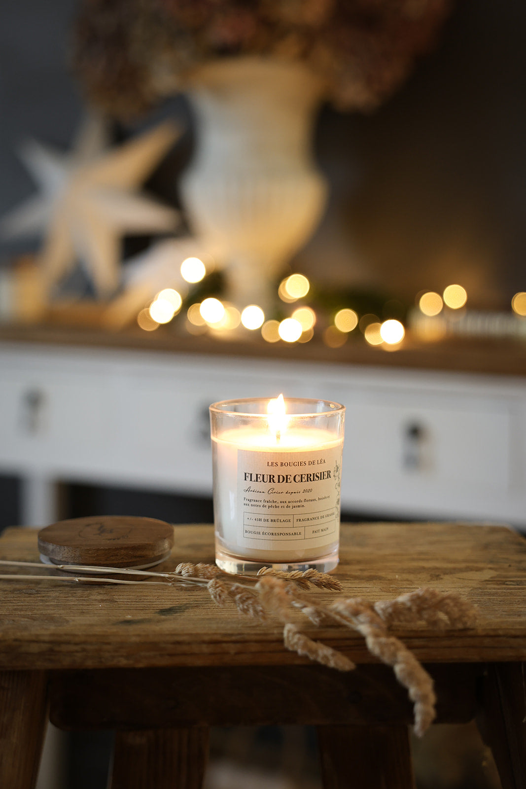 Bougie parfumée artisanale Les Bougies de Léa Fleur de Cerisier allumée, posée sur un tabouret en bois et branche de blé séché et guirlandes lumineuses et étoile déco en fond.
