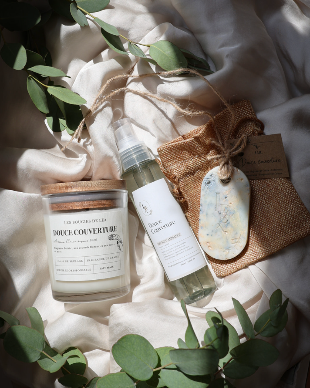 Bougie parfumée, brume d'ambiance et suspension végétale parfumée LBL Les Bougies de Léa de la senteur Douce couverture, sur un drapé blanc satiné, à la lumière naturelle du soleil, entourés de feuilles d'eucalptus.