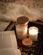 Bougie parfumée Douce couverture Les Bougies de Léa sur une table accompagnée de livres ouverts et plaid ou une couverture en fond, et d'une autre petite bougie parfumée Les Bougies de Léa allumée pour une ambiance chaleureuse et automnale.