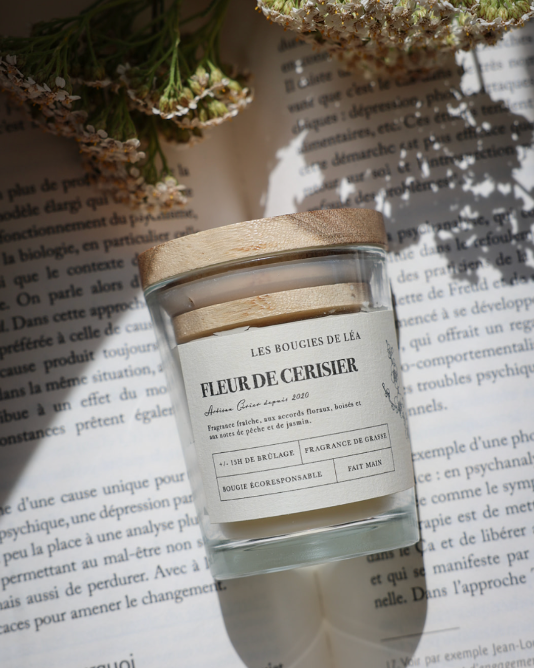 Bougie artisanale parfumée Fleur de Cerisier Les Bougies de Léa sur un livre ouvert et fleurs blanches, avec rayons de soleil
