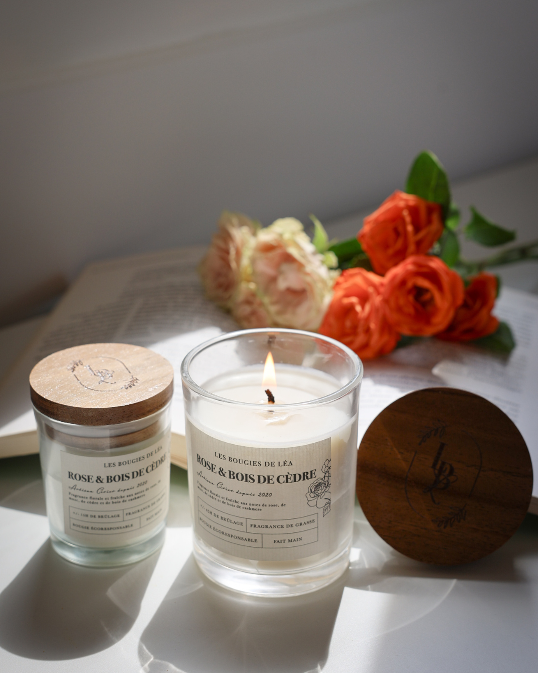 Bougies artisanales parfumées Rose & Bois de Cèdre Les Bougies de Léa allumée, sur une table avec un livre ouvert et des roses, à la lumière du soleil.