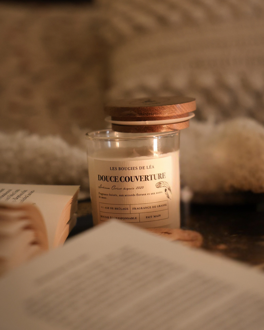 Bougie parfumée Douce couverture Les Bougies de Léa sur une table accompagnée de livre ouverts et coussin blanc en fond.