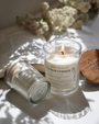 Bougie parfumée Fleur de Cerisier Les Bougies de Léa allumée grand et petit format avec étiquette en papier recyclé et couvercle en bois, posées sur une table blanche, au rayons du soleil avec fleurs fleurs et livre en fond.