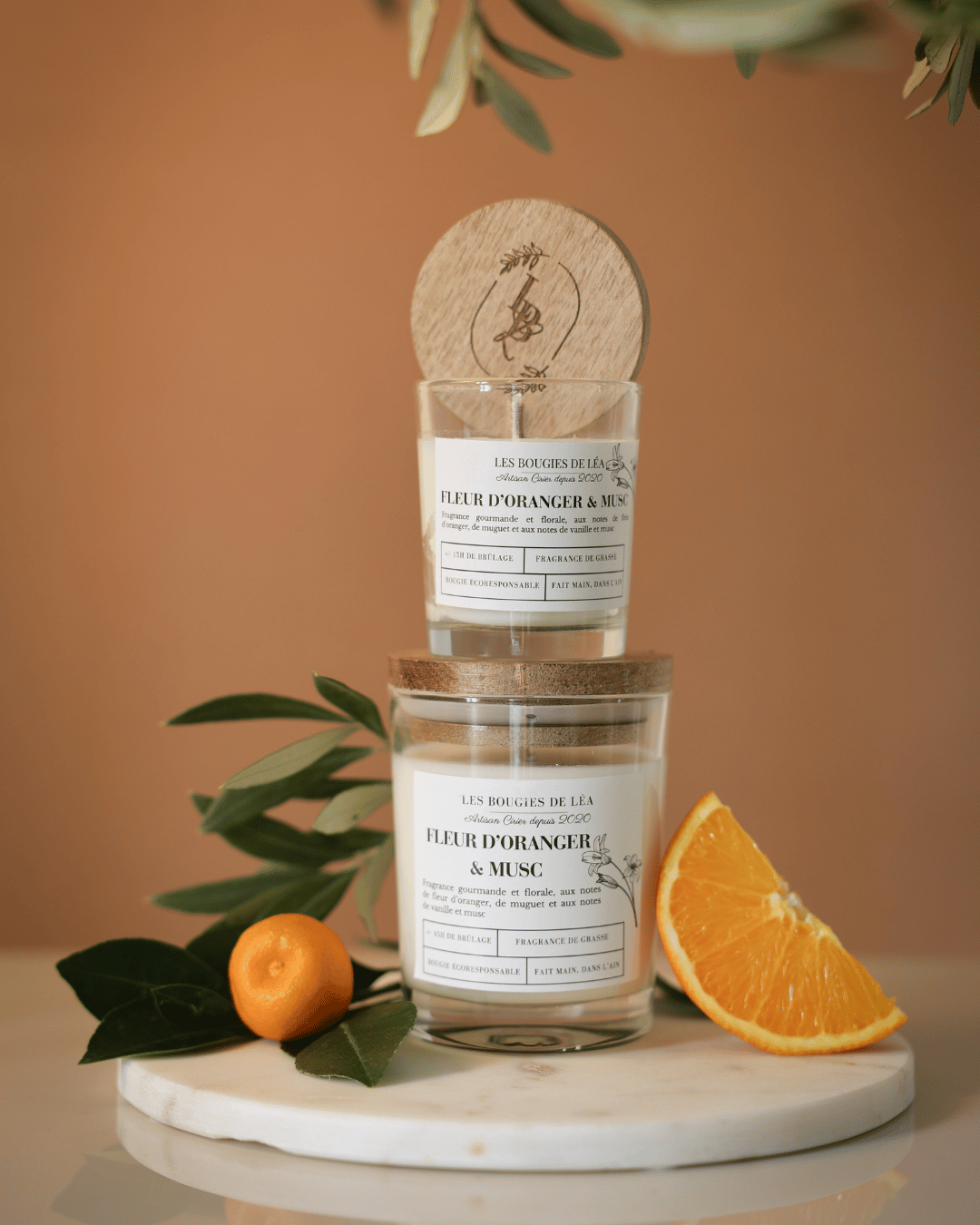 Bougies artisanales parfumées Fleur d'oranger & Musc, petit et grand modèle, Les Bougies de Léa posée sur un support en marbre et fond beige, accompagnées d'oranges et de feuilles d'olivier pour une ambiance provençale.