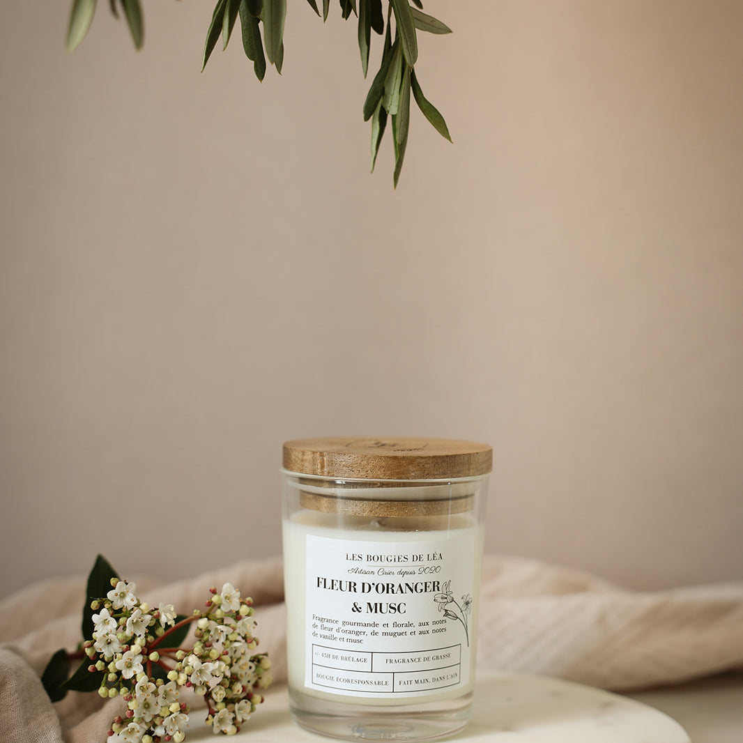 Bougie artisanale parfumée Fleur d'oranger & Musc Les Bougies de Léa allumée avec un drap en lin en arrière plan, accompagnée de fleurs blanches et feuilles d'olivier.