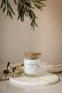 Bougie artisanale parfumée Fleur d'oranger & Musc Les Bougies de Léa allumée avec un drap en lin en arrière plan, accompagnée de fleurs blanches et feuilles d'olivier.
