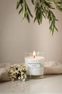 Bougie artisanale parfumée Fleur d'oranger & Musc Les Bougies de Léa allumée avec un drap en lin en arrière plan, accompagnée de fleurs blanches et feuilles d'olivier.