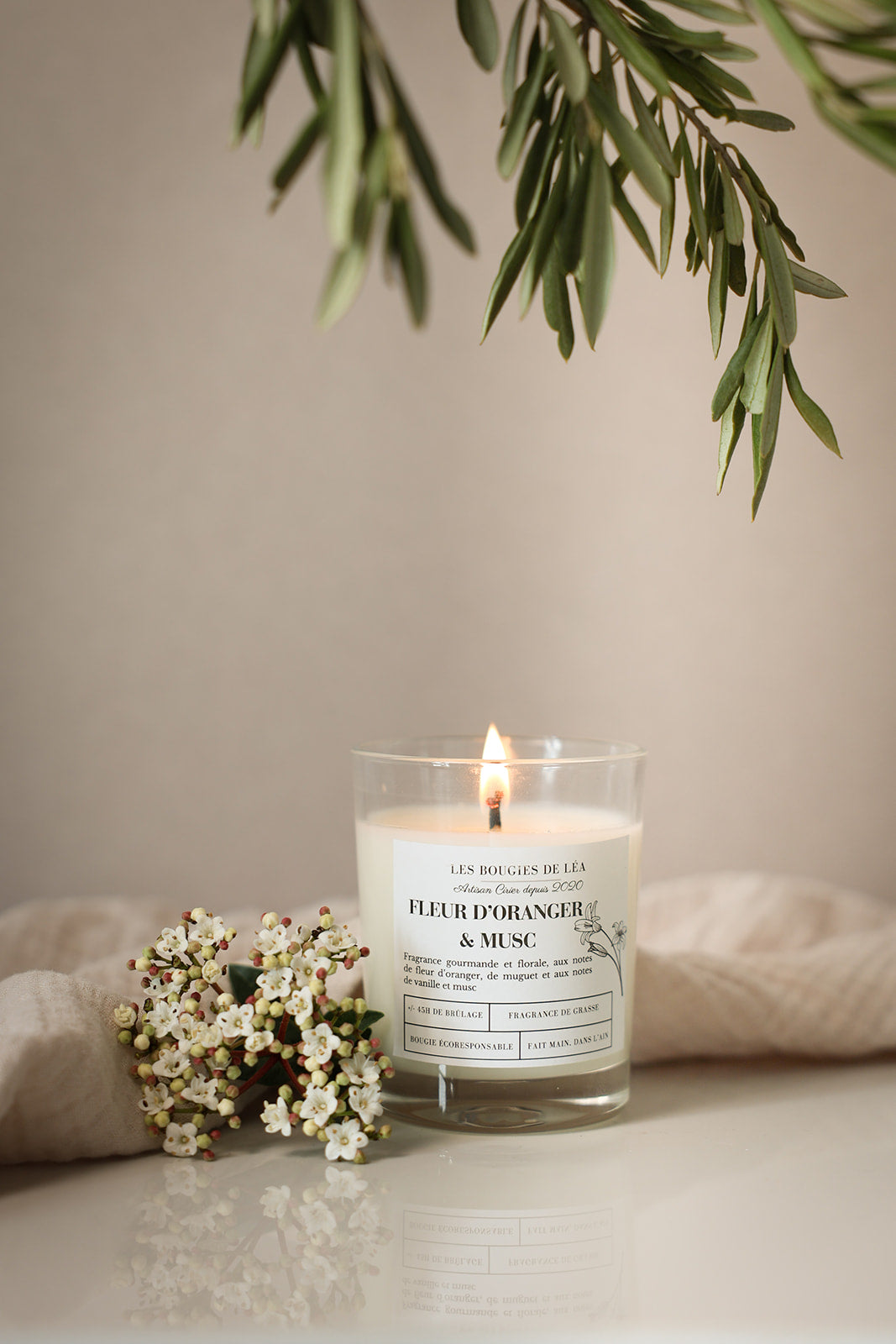 Bougie artisanale parfumée Fleur d'oranger & Musc Les Bougies de Léa allumée avec un drap en lin en arrière plan, accompagnée de fleurs blanches et feuilles d'olivier.