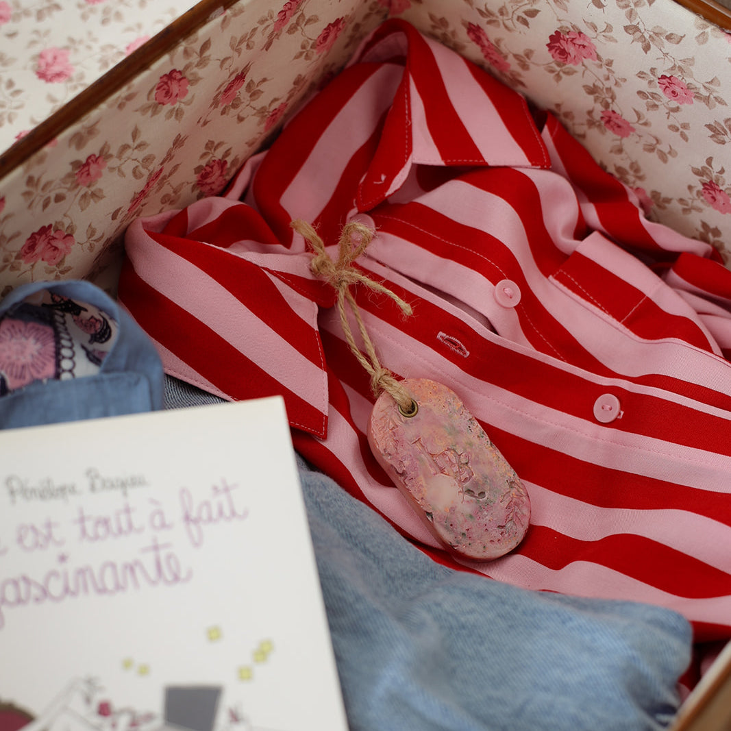 Suspension artisanale parfumée LBL Pêche de vigne dans un valise sur un chemisier rose et rouge à rayure pour parfumer les vêtements, accompagné d'un livre pour les vacances.