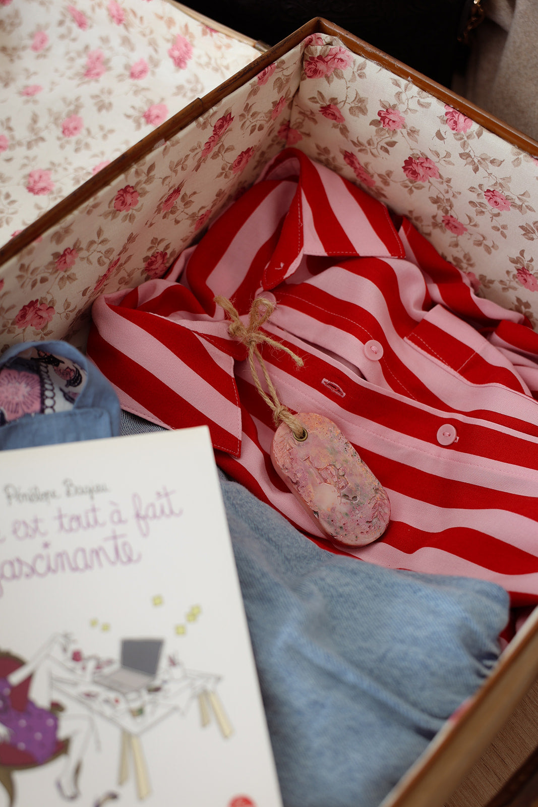 Suspension artisanale parfumée LBL Pêche de vigne dans un valise sur un chemisier rose et rouge à rayure pour parfumer les vêtements, accompagné d'un livre pour les vacances.