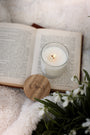 Bougie Poudre de riz posée sur un livre ouvert, entourée d'un plaid blanc et de fleurs, ambiance lecture, cocooning et détente.