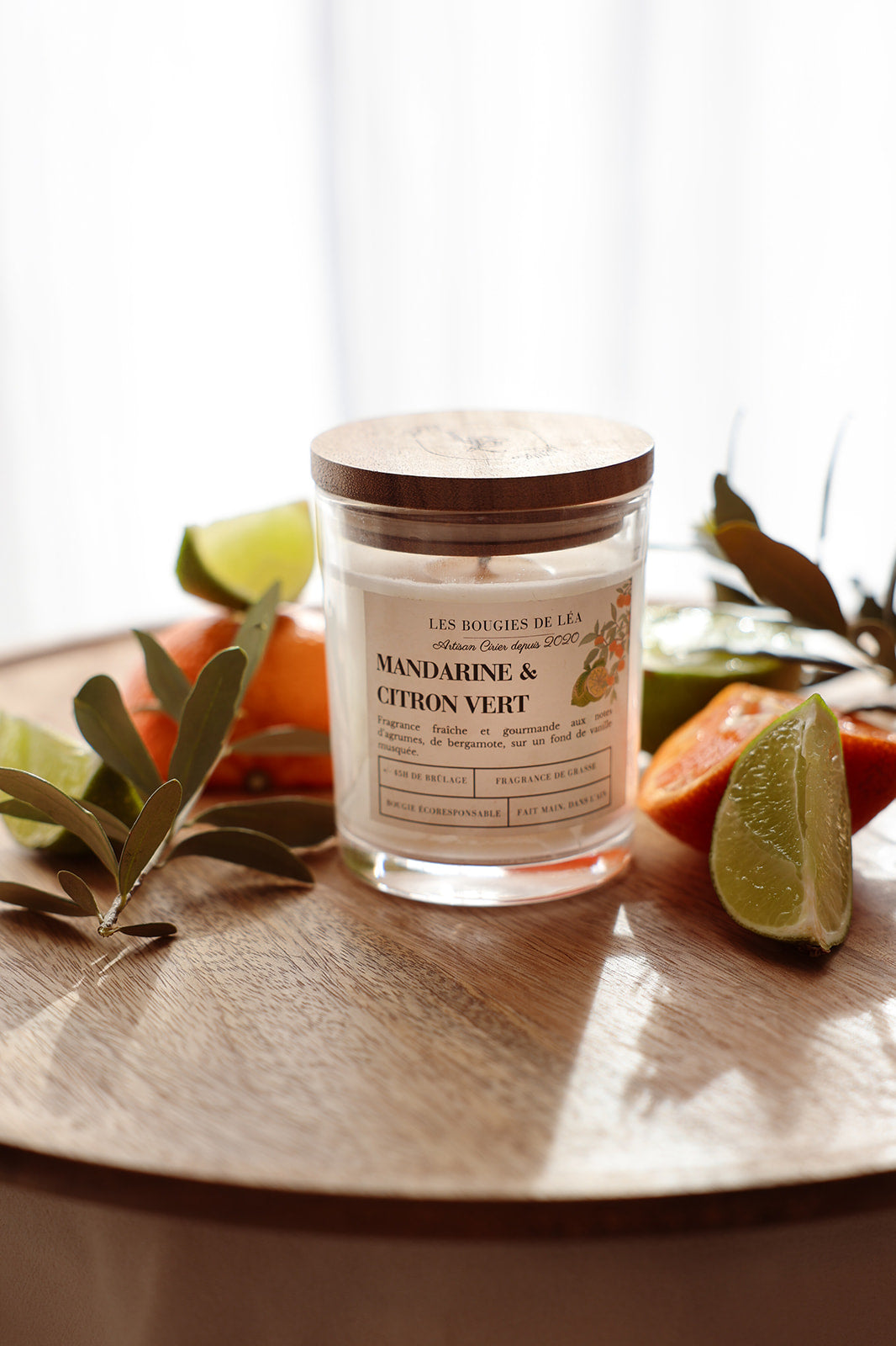 Bougie artisanale parfumée Mandarine & Citron vert Bergamote Les Bougies de Léa couvercle en bois d'acacia, sur une table en bois accompagnée d'agrumes, oranges, citron vert et feuilles d'olivier.