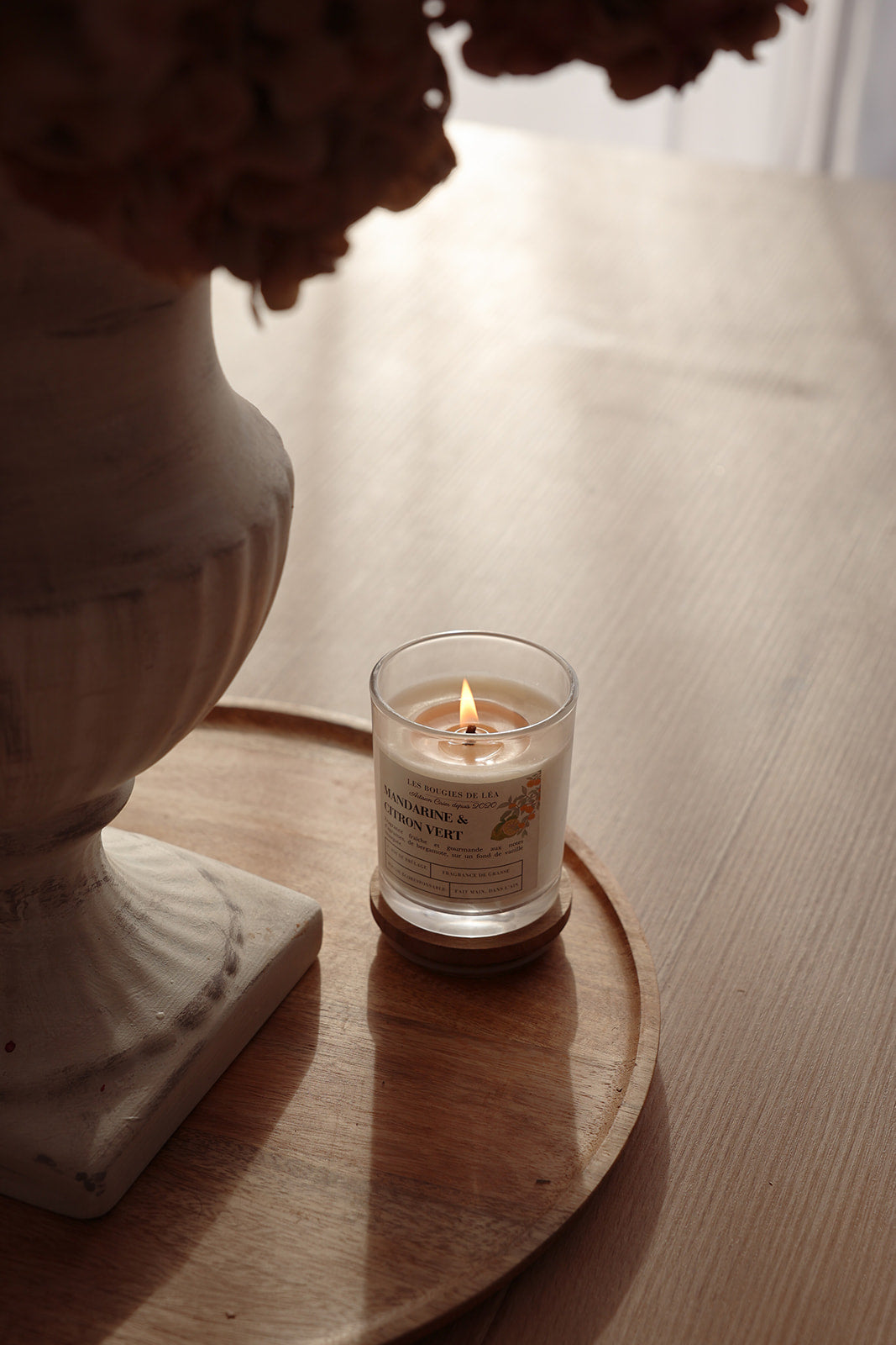 Bougie artisanale parfumée Mandarine citron vert Bergamote Les Bougies de Léa allumée, sur une table et support en bois, à côté d'un vase de fleurs séchées.