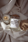 Bougie artisanale parfumée Les Bougies de Léa Pêche de vigne sur un textile de maison blanc satin, couvercle en bois d'acacia, accompagnée d'une petite bougie allumée.