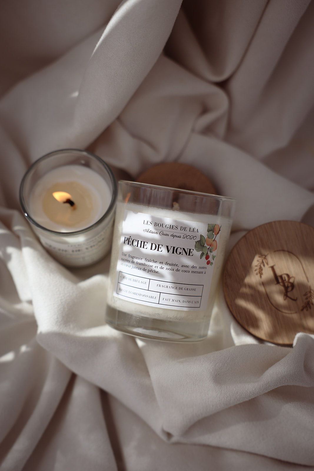 Bougie artisanale parfumée Les Bougies de Léa Pêche de vigne sur un textile de maison blanc satin, couvercle en bois d'acacia, accompagnée d'une petite bougie allumée.