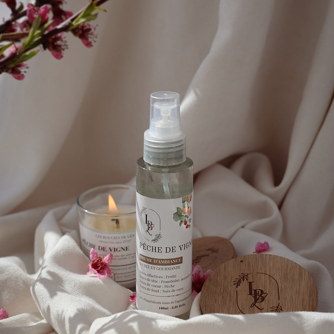 Brume d'ambiance parfumée végétale LBL Pêche de vigne sur un drap textile de maison blanc satin, accompagné d'une bougie parfumée artisanale Les Bougies de Léa allumée Pêche de Vigne, entouré de Fleurs de Cerisier.