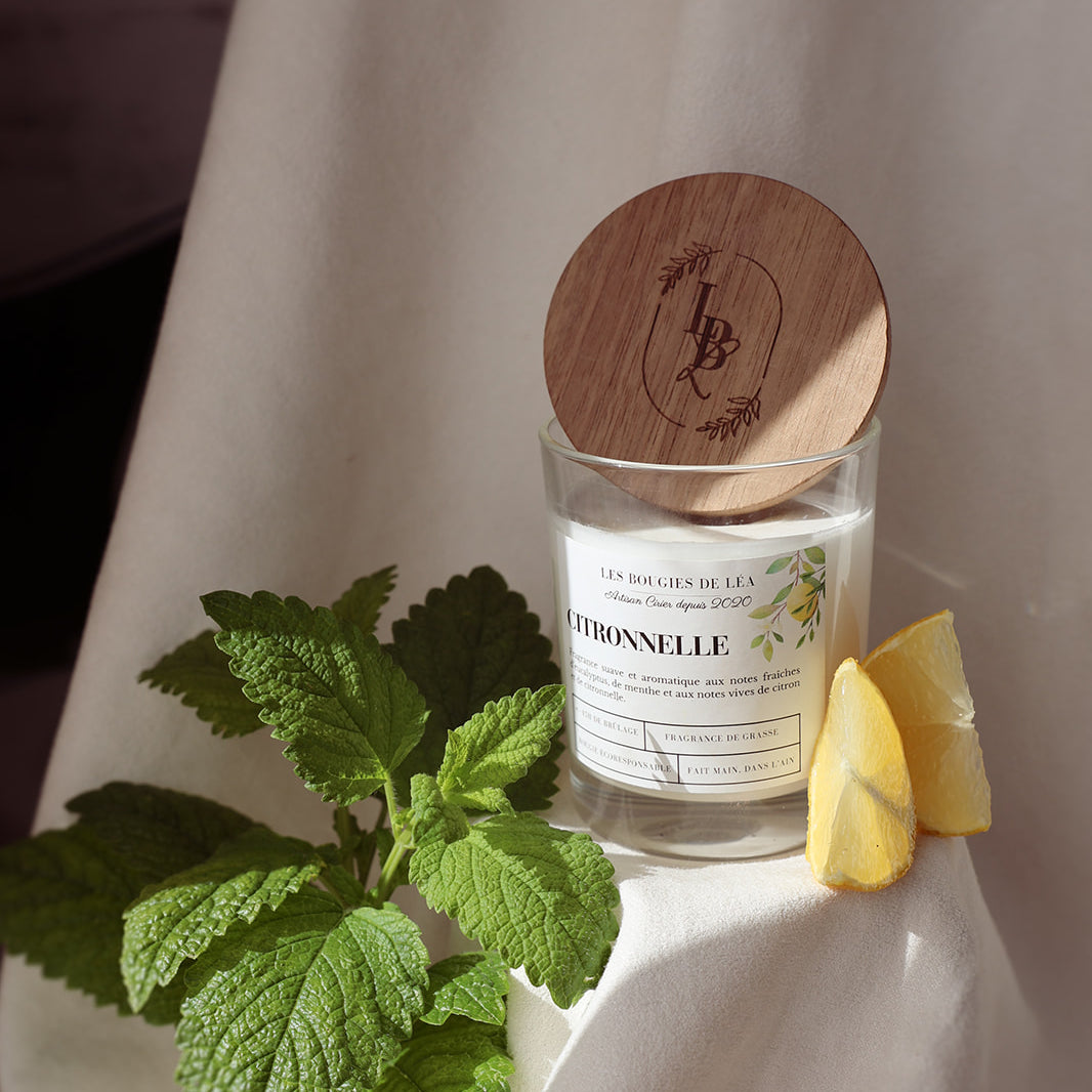 Bougie artisanale parfumée Les Bougies de Léa Citronnelle avec couvercle en bois d'acacia, sur un drap blanc ou une nappe, accompagnée de tranches de citron jaune et feuilles de menthe.