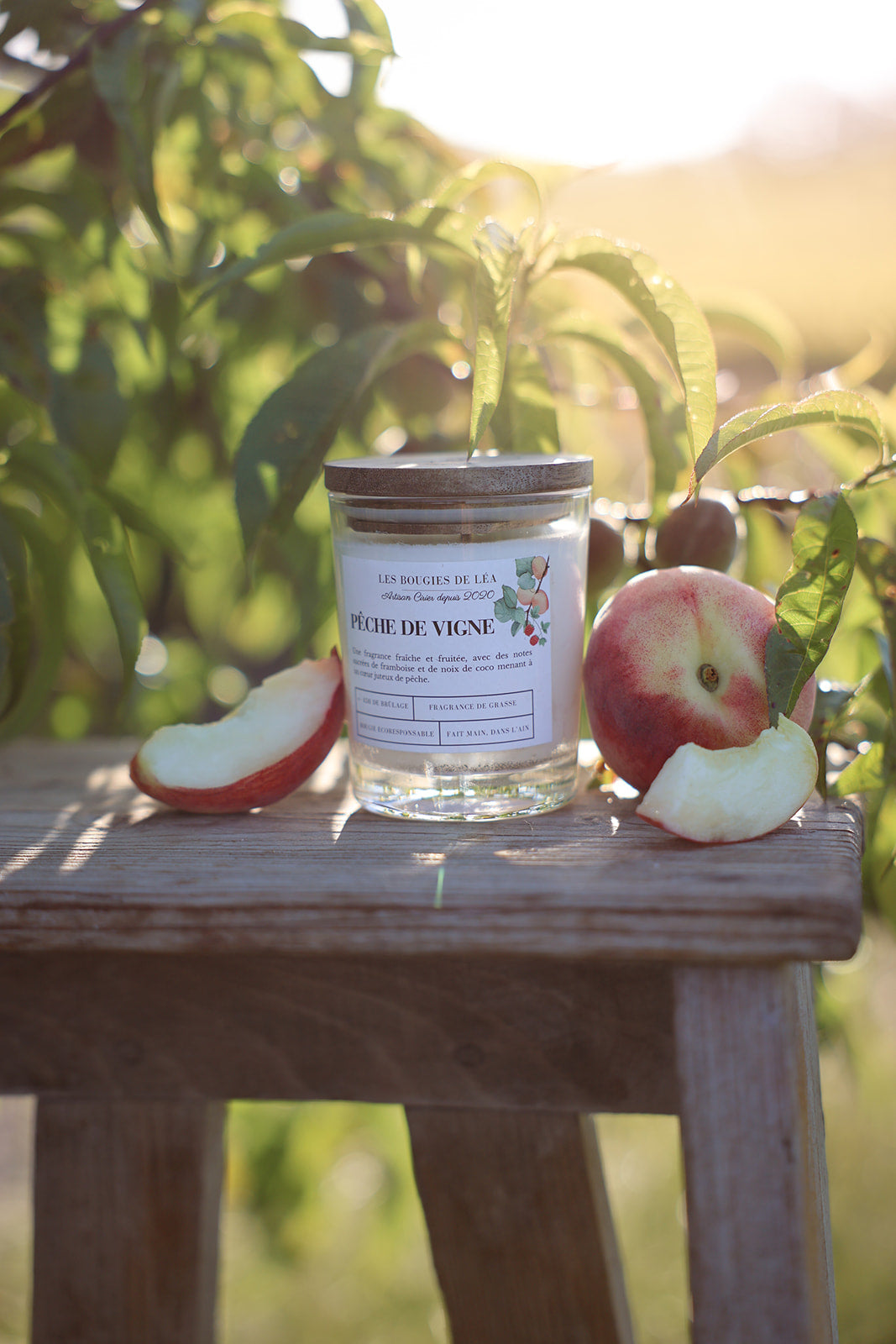 Bougie artisanale parfumée Les Bougies de Léa Pêche de vigne avec son couvercle en bois d'Acacia, posée sur un tabouret en bois, dans les vignes à la lumière du soleil accompagnée de pêches juteuses.