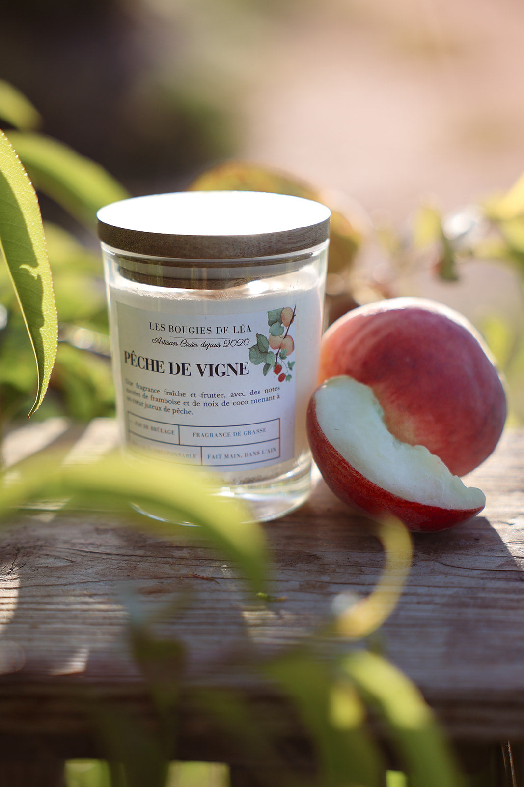 Bougie artisanale parfumée Les Bougies de Léa Pêche de vigne avec son couvercle en bois d'Acacia, posée sur un support en bois, dans les vignes à la lumière du soleil accompagnée de pêches juteuses.