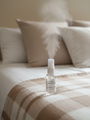 Flacon testeur brume d'ambiance Douce couverture LBL sur le draps d'un lit beige et marron avec oreillers et coussins en arrière plan.